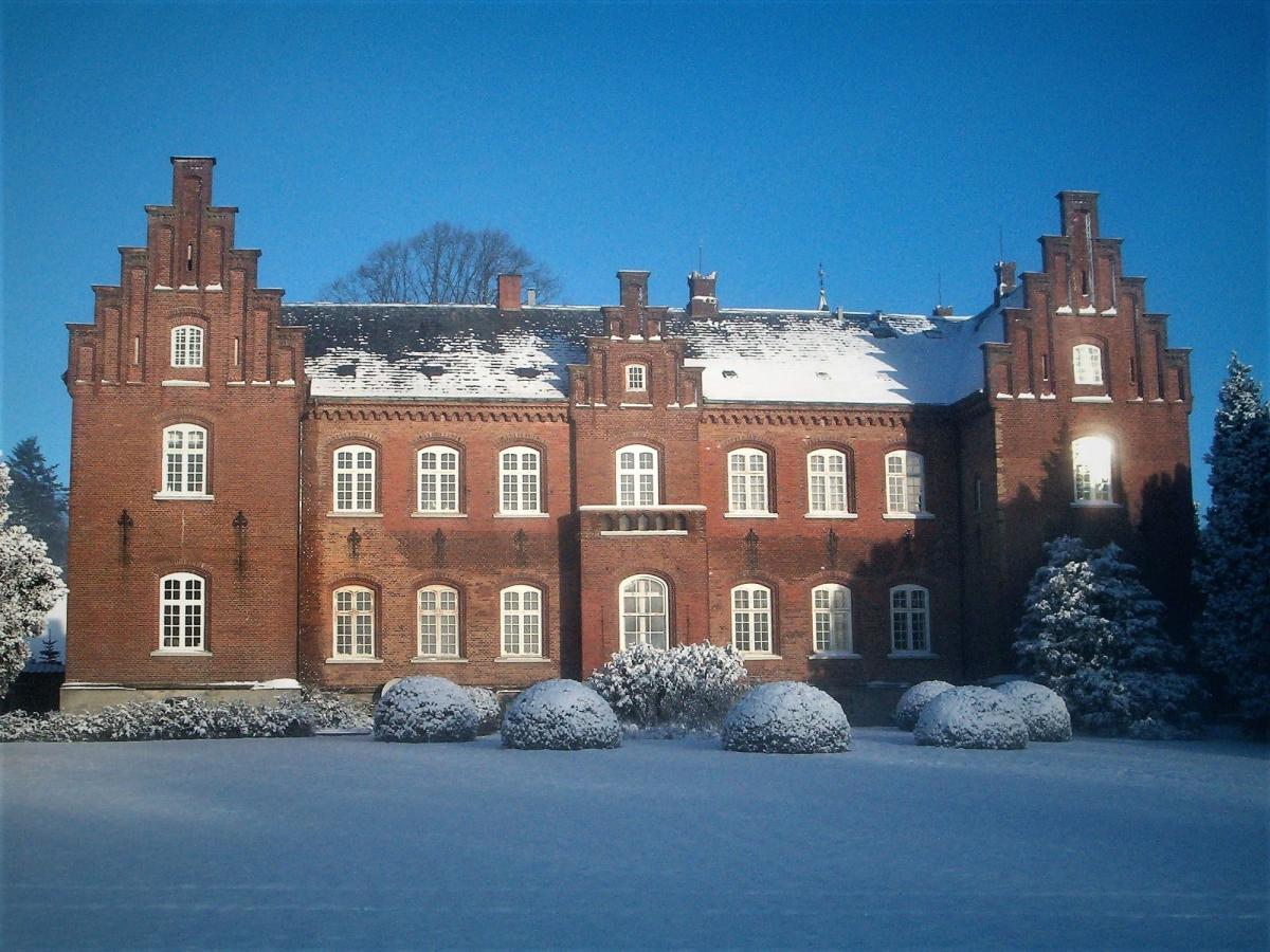 En rød slotsbygning med hvide vinduesrammer lyser op i et snedækket landskab. På det sorte tag ligger sne, og foran slottet er græsplænen og buskene dækket af sne.