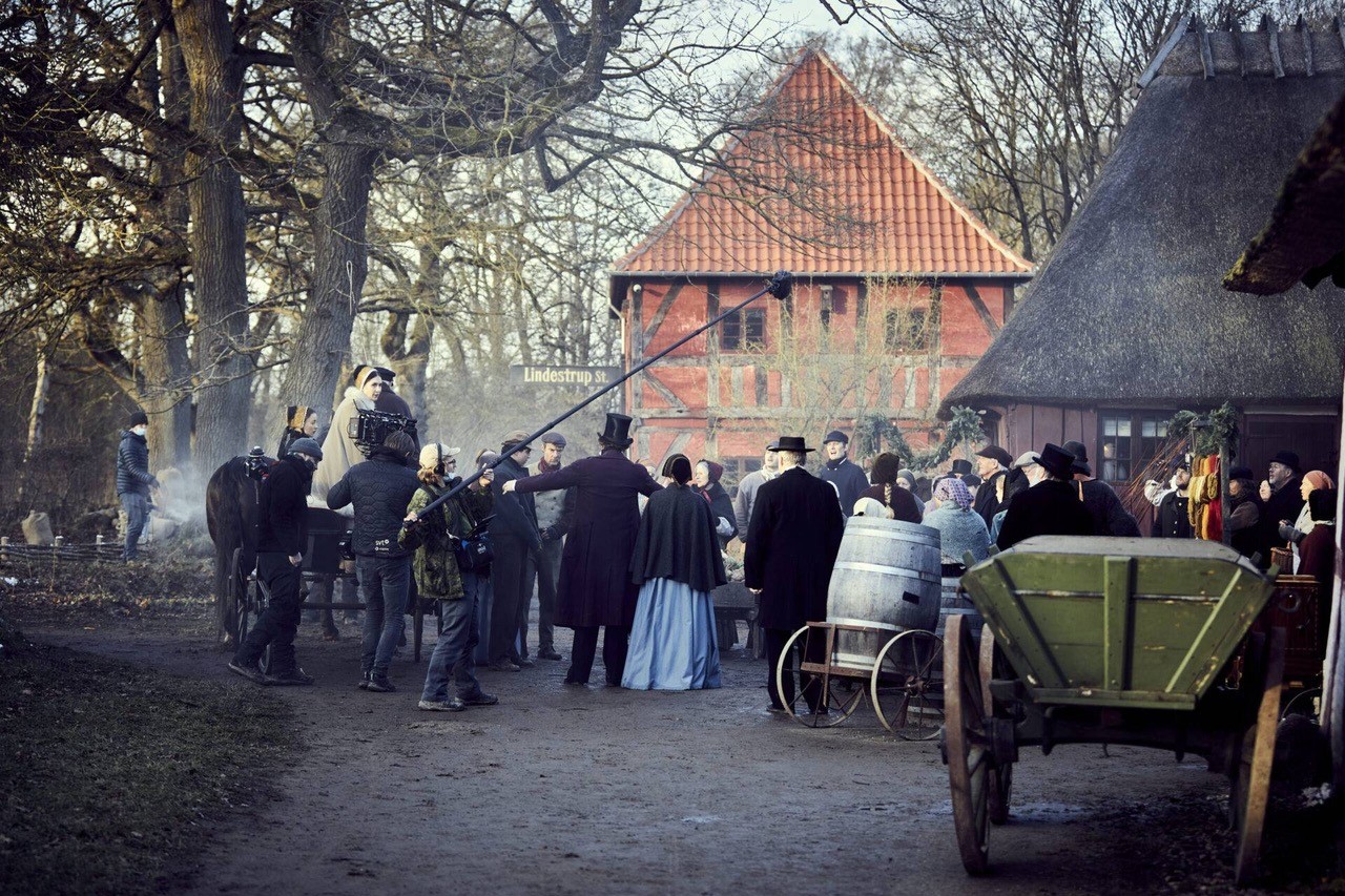 Et billede fra en filmoptagelse hvor der står både en mand med mikrofonstang og en mand med et kamera. De filmer en masse mennesker på en grusvej som står i gammeldags tøj. De står mellem to bindingsværkshuse. Til højre på billedet står en grøn hestekærre.