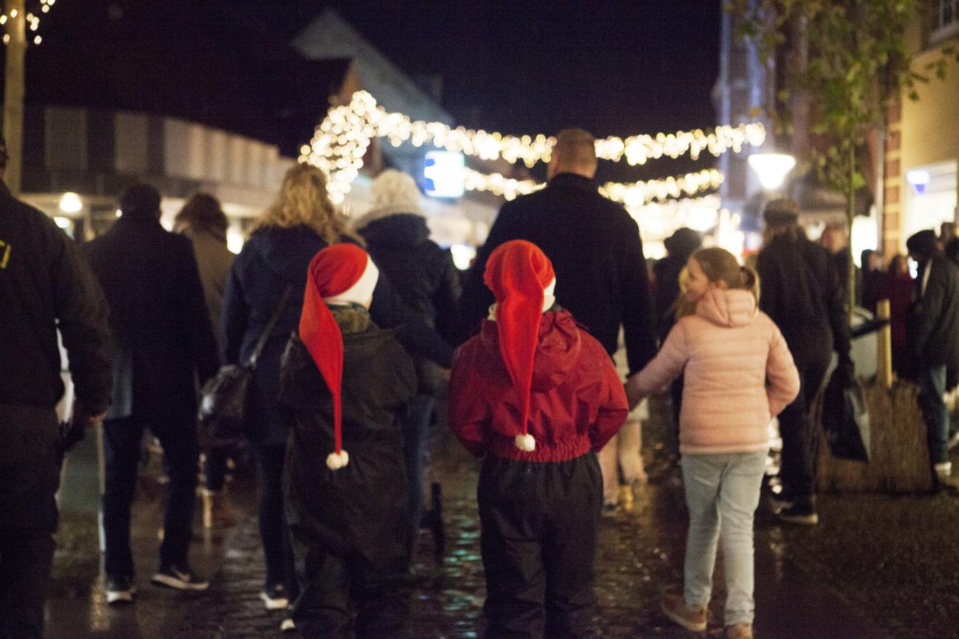 En gågade oplyst af julelys. Der går en masse mennesker men i forgrunden går tre piger. Til højre går en pige i lyse cowboybukser og en lyserød jakke. De to piger til højre har begge flyverdragter på og en lang nissehue.