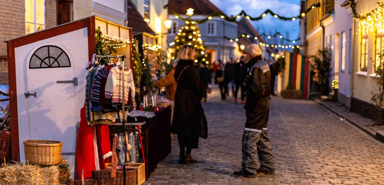 En brostensgade. Til højre er en række af små farvede huse. Til venstre er en julemarkedsbod sat op. Der er flettede kurve, børne striksweatres og julelys. Foran boden står en mand og kvinde og taler. I baggrunden kan man spotte flere slørede mennesker og boder.