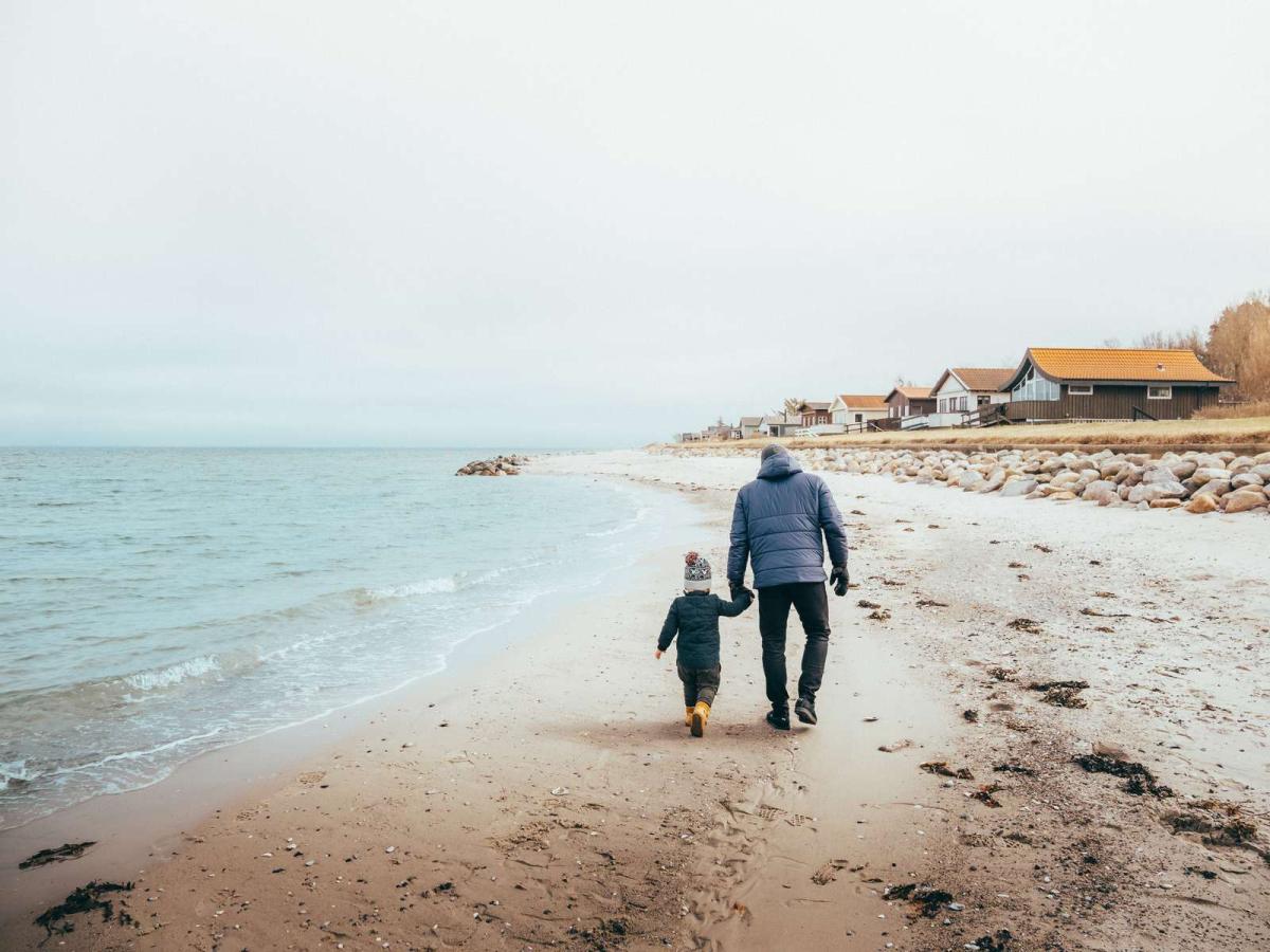 Far og søn har varmt tøj på, og går hånd i hånd på en strand med sommerhuse i baggrunden