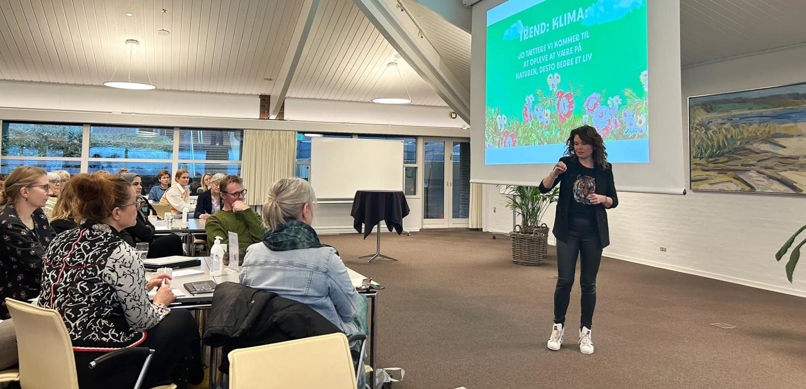 Julia Lahme på workshoppen i Svendborg om adfærdsdesign og bæredygtighed. Hun står foran en skærm med powerpoint på. Til venstre i billedet sidder der mennesker rundt om firkantede borde og lytter.