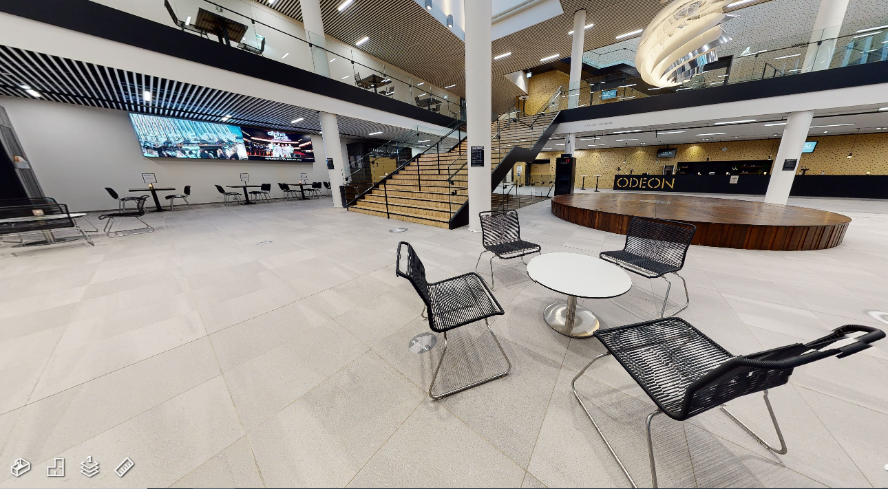 Stor tom sal med borde, stole, spotlamper og reklamebilleder.  I forgrunden ses fire sorte stole samlet omkring et lille hvidt bord.