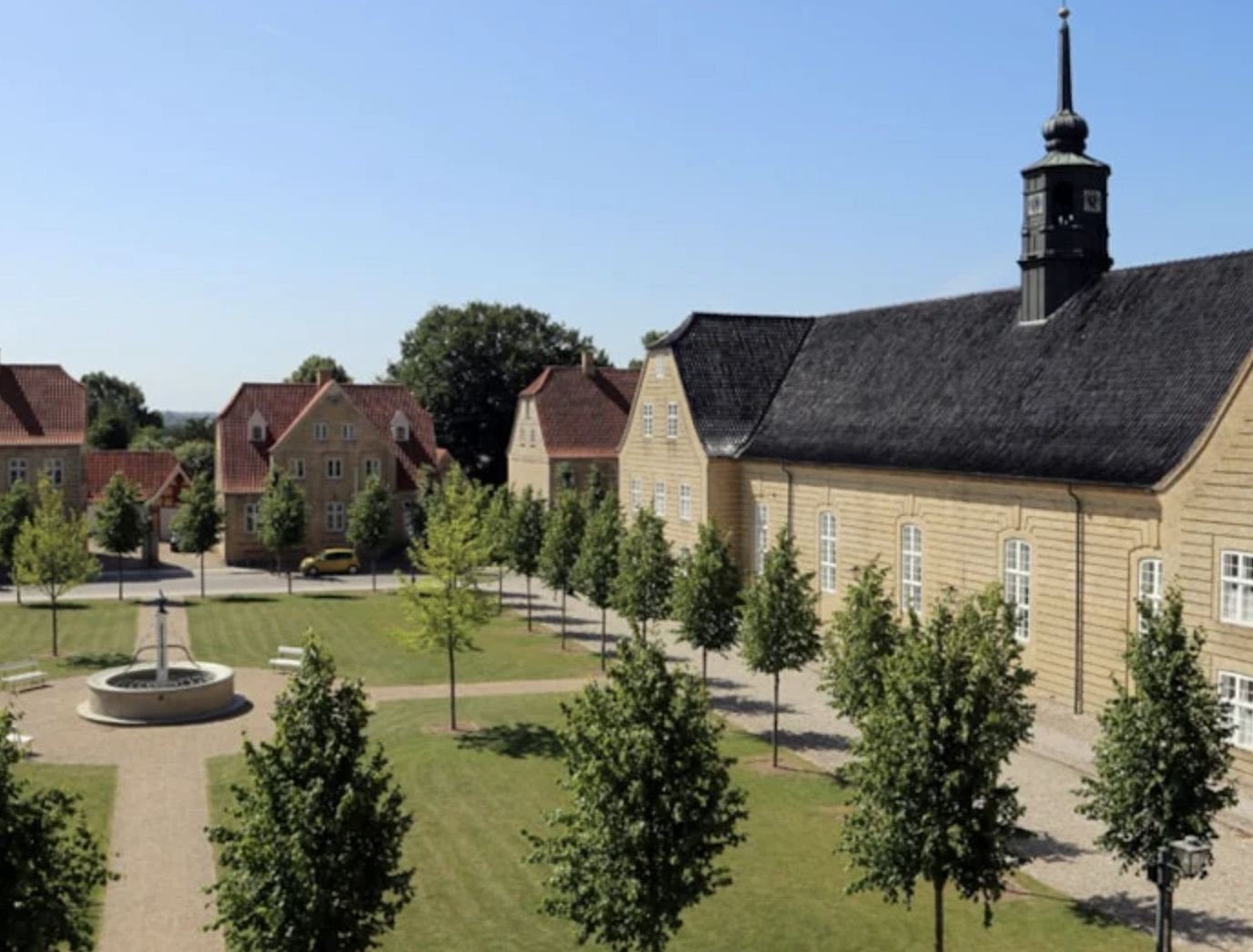 Søstrehuset i byen Christiansfeld. Gul gårdbygning med sort tag. Grøn plæne med symmetriske grønne træer foran.