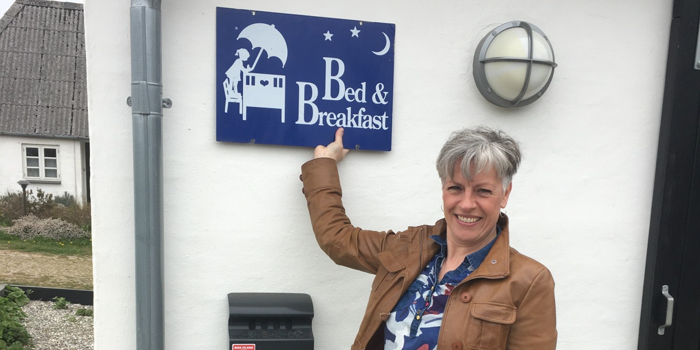 Kamilla fra Aasesminde B&B peger på et blåt Ole Lukøje-Bed & Breakfast skilt på en hvid mur