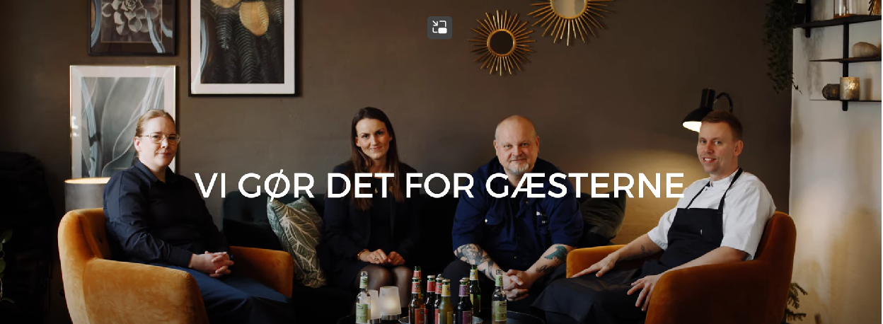 En kvinde og tre mænd sidder i lænestole rundt om et sofabord. På billedet står teksten "Vi gør det for gæsterne"