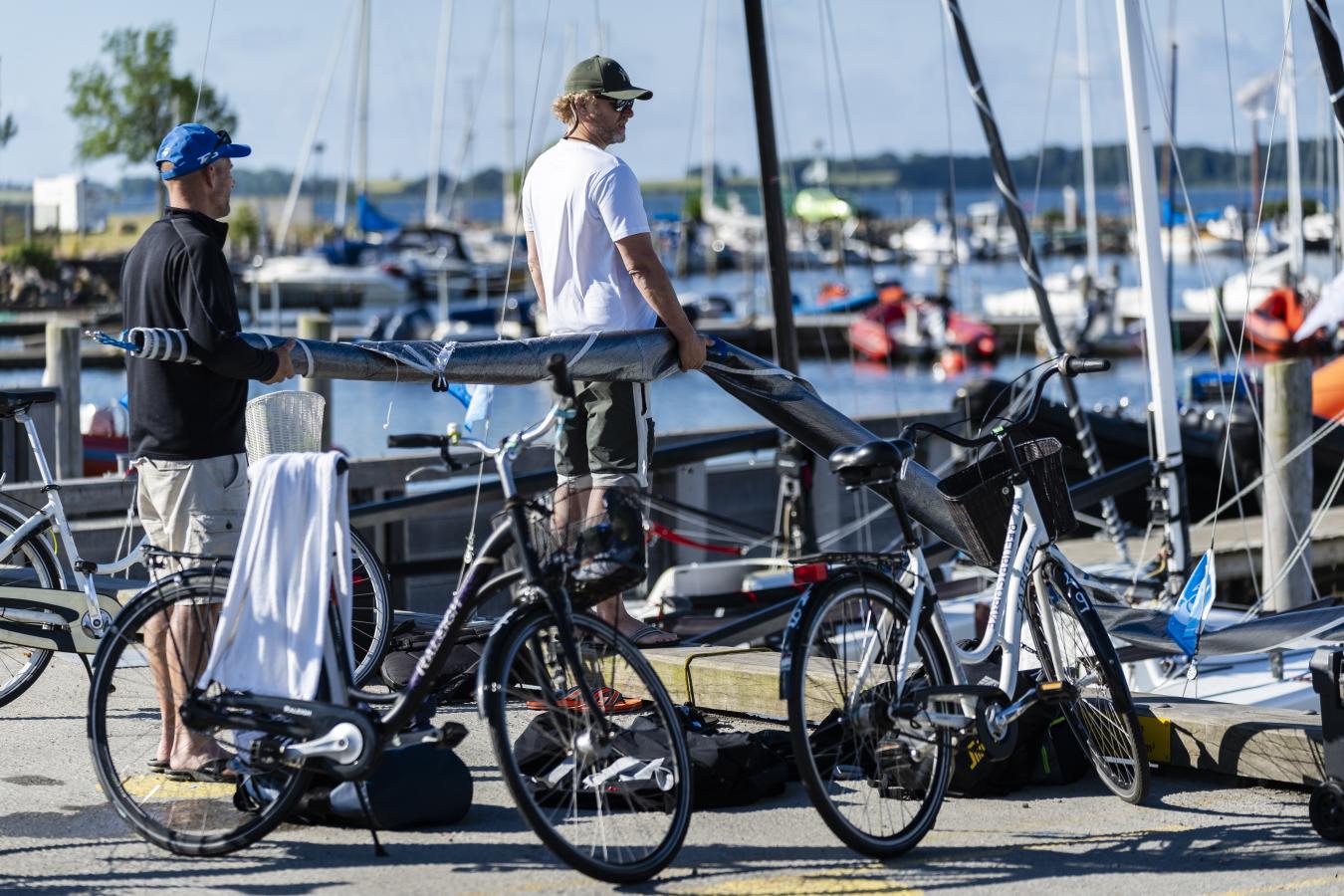 To cykler står parkeret på Middelfart Marina. I baggrunden er to sejlere i gang med at bære et sammenrullet sejl ned på en sejlbåd. 