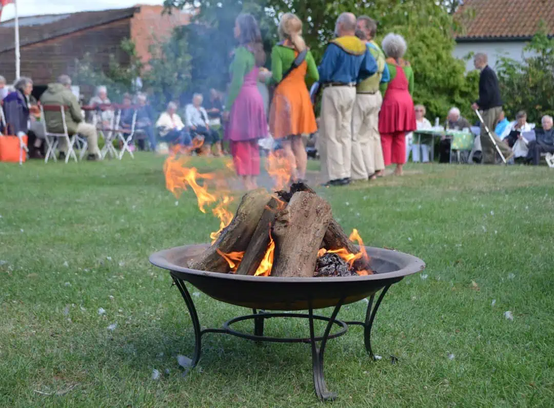 I forgrunden ses et bålfad med et bål og i baggrunden ses en gruppe mennesker i farverigt tøj.