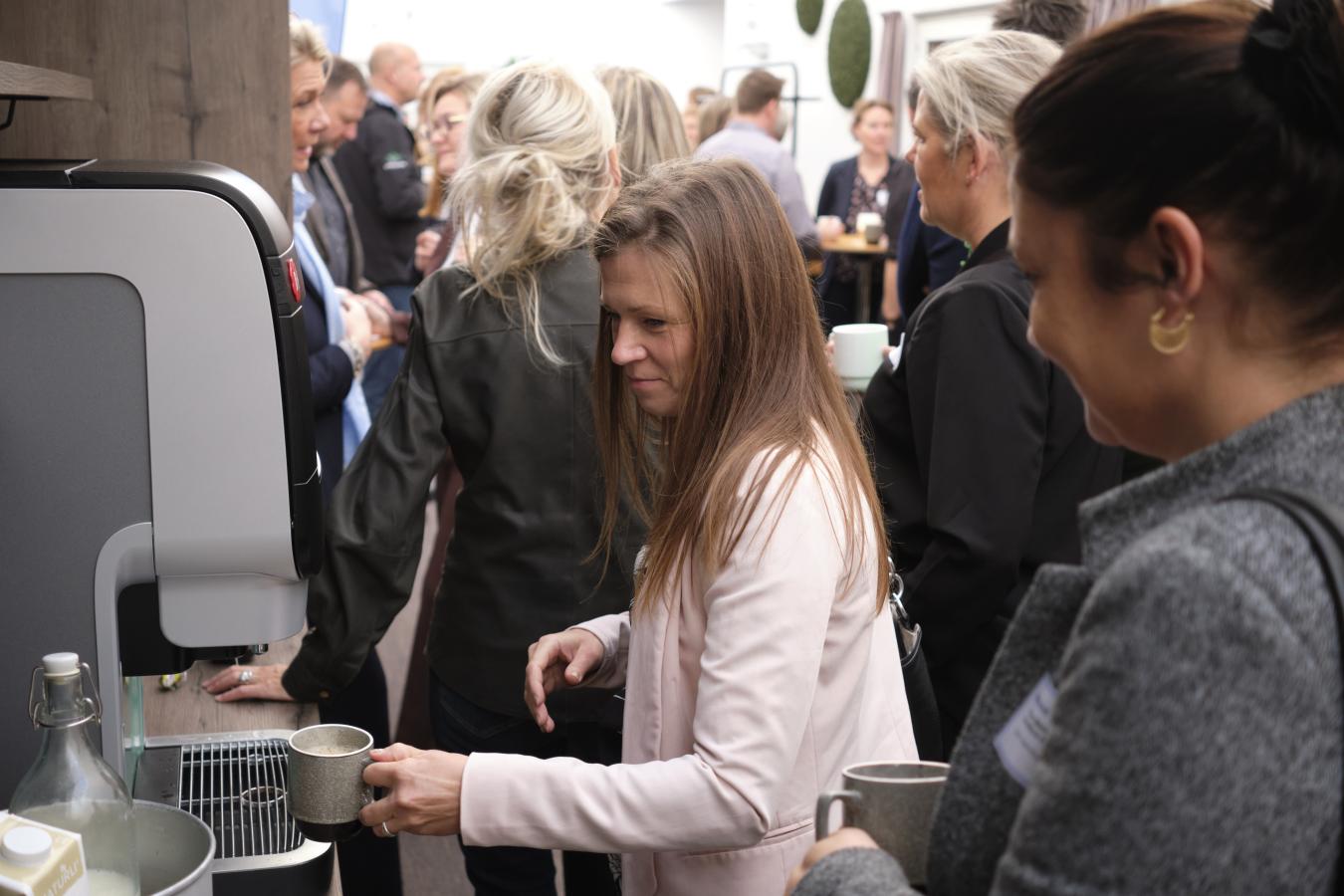 Kvinde i lyserød blazer tapper kaffe i kaffeautomaten på en konference, mens en anden kvinde venter på hendes tur. Mange mennesker i baggrunden.