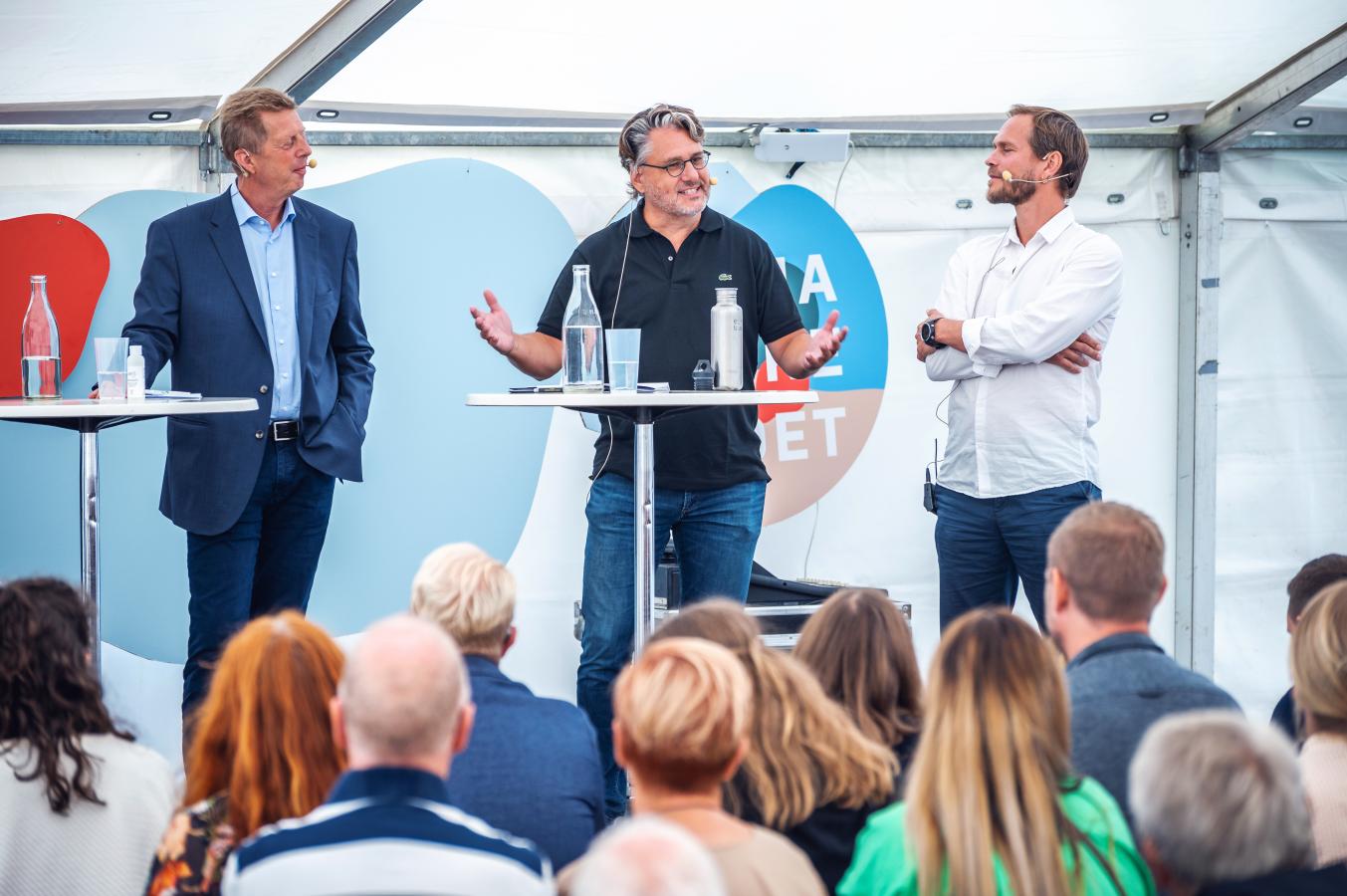 Tre mænd står på en scene og debatterer ved nogle høre caféborde med vandflasker på. I forgrumden ses baghovederne af tilhørerne.