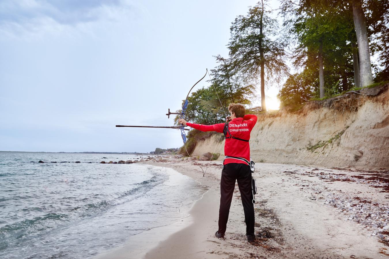 Bueskytte står på strand med ryggen til med buen spændt og sigter ud over vandet.