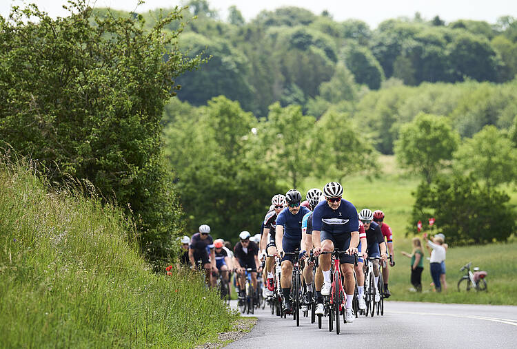En flok cykelryttere på vej op ad bakke på asfaltvej med grønne marker og træer omkring dem. Et par tilskuere hepper i vejkanten.
