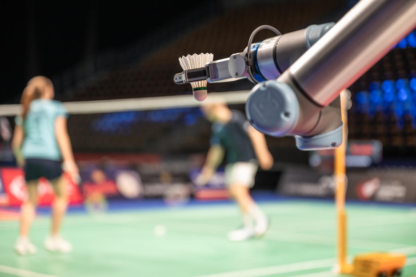 Robotarm med badmintonfjerbold. I baggrunden ser to spillere på en badmintonbane.