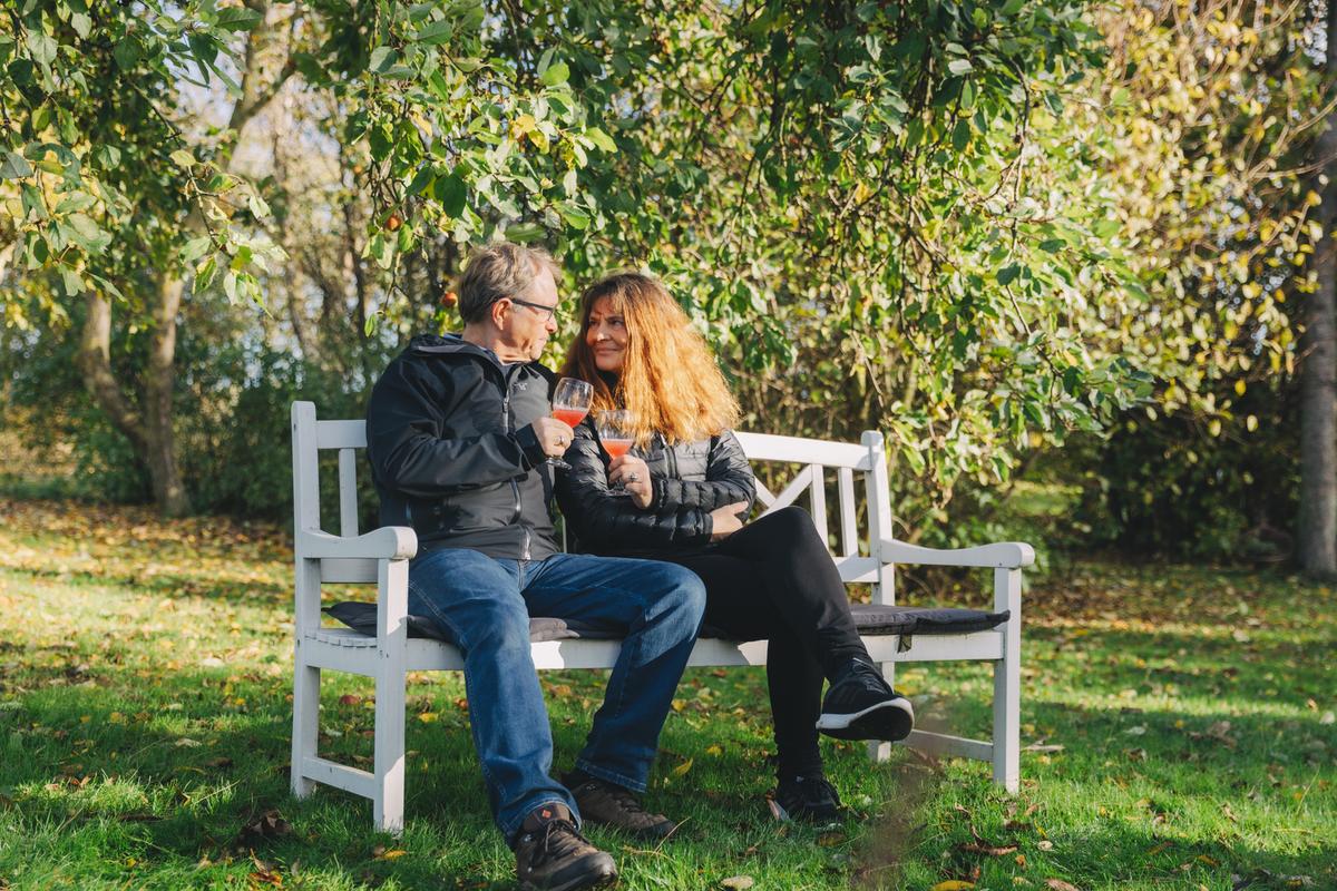 Et par sidder og skåler på en hvid bænk under et træ i en grøn have