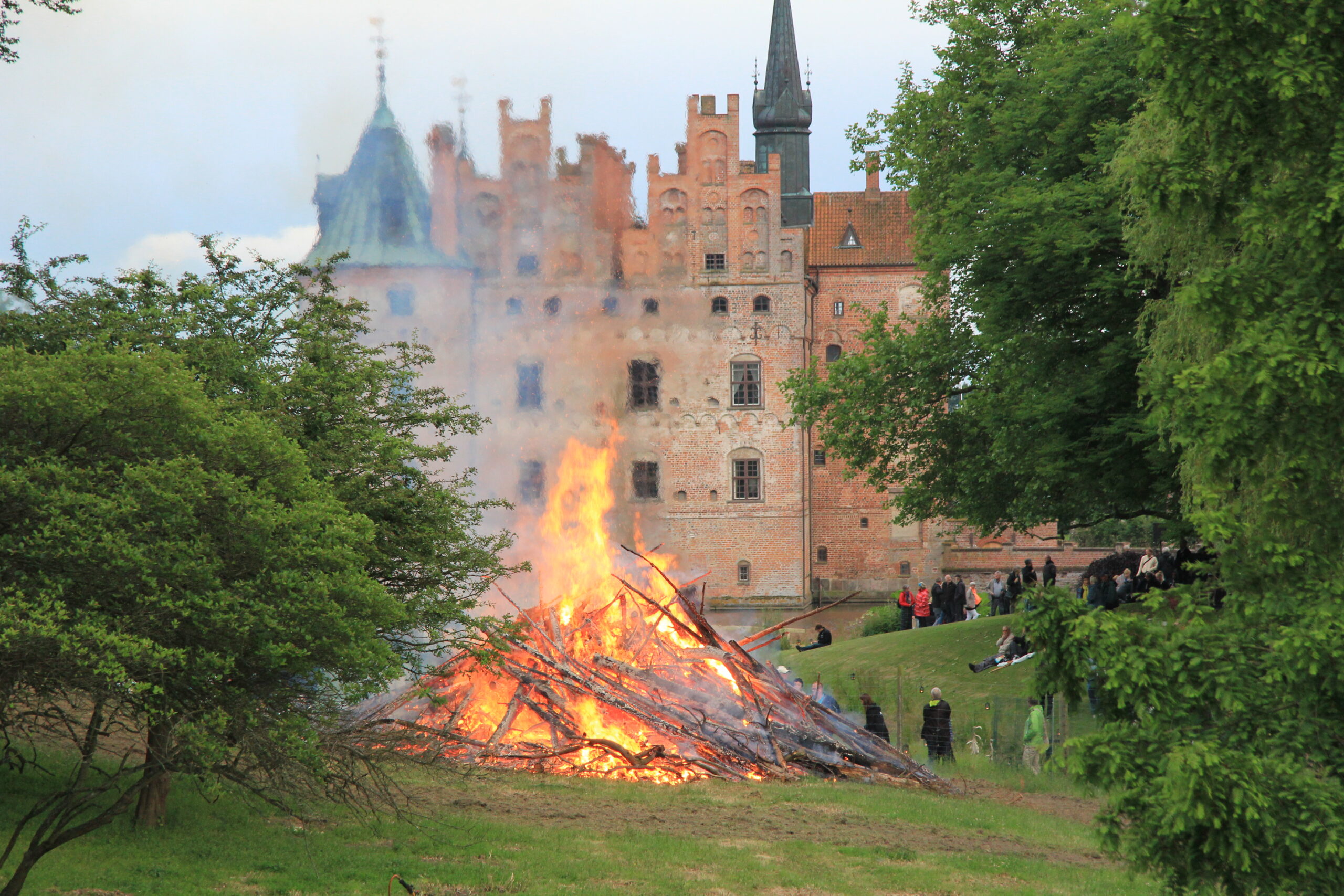 Egeskov i baggrunden, et skt hans bål i forgrunden omkranset af grønne buskadser og træer