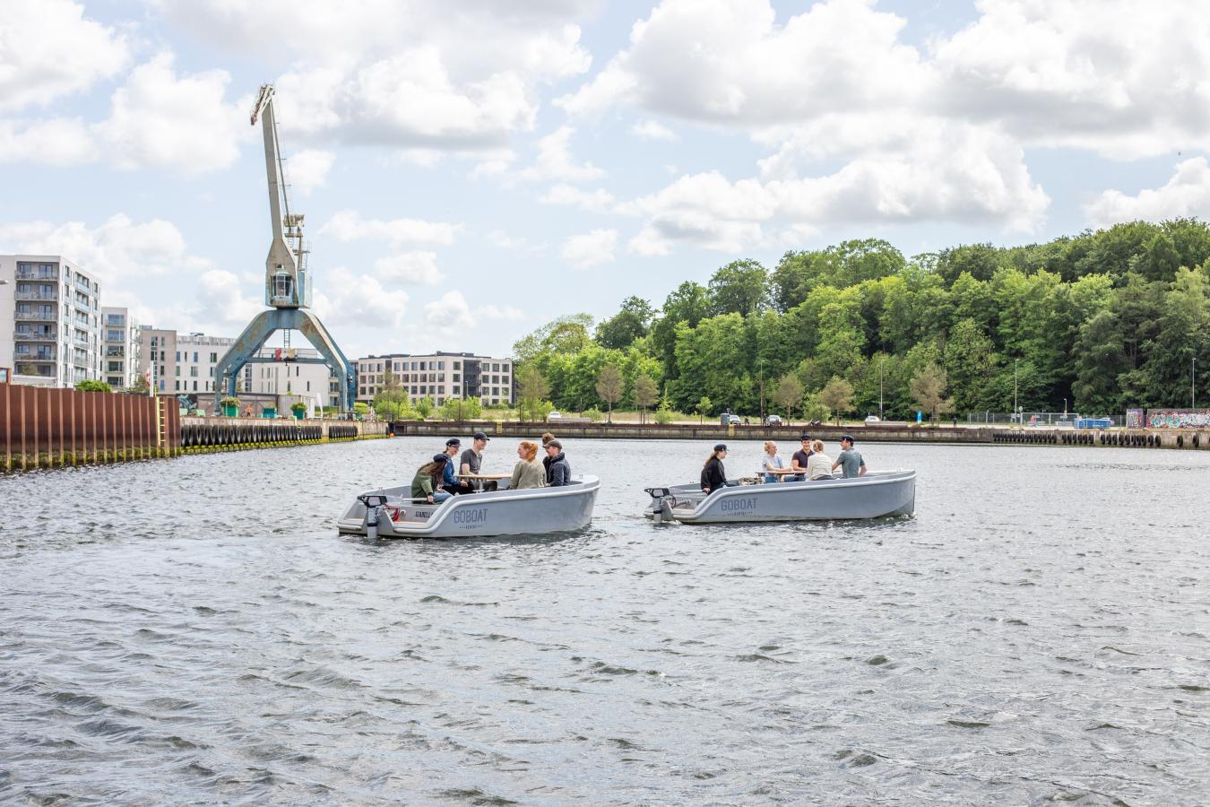 To GoBoat-både på vandet ved Odense Havn med grønne træer og en stor kran i baggrunden