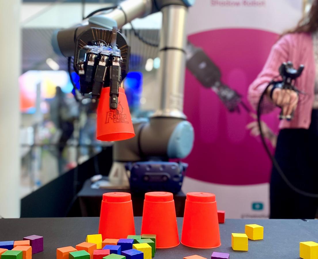 Robotarm løfter et rødt bæger ved et bord med flere røde bægre og klodser i forskellige farver