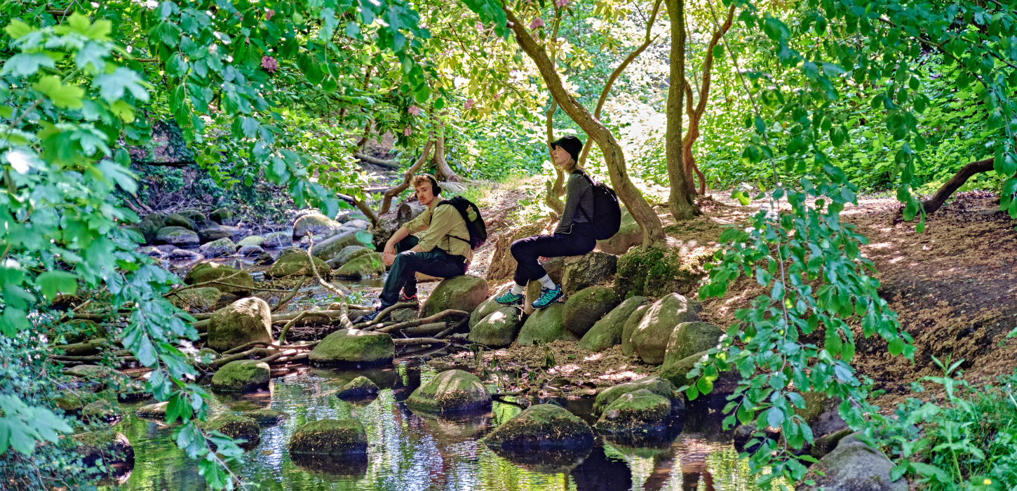 To vandrere sidder på et stenleje ved siden af en lille å i en grøn skov