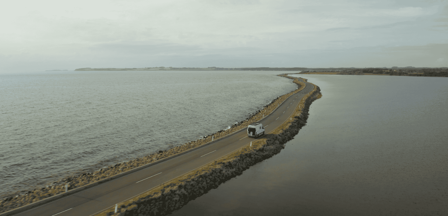 A white motorhome driving on a narrow dam with ocean to both sides