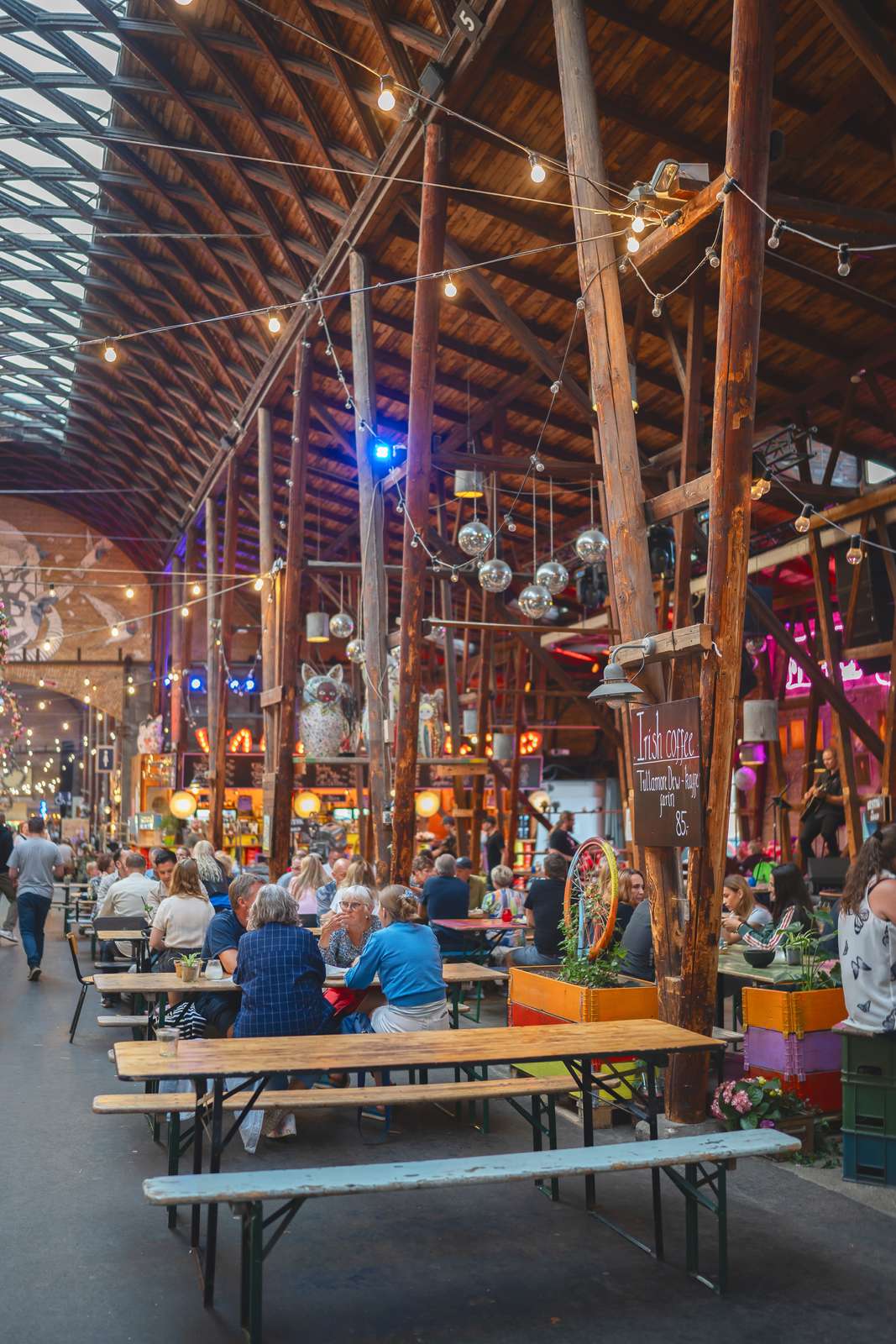 Et gammelt pakhus med træstolper med madboder, bord og bænkesæt, hyggebelysning og mennesker der hygger sig 