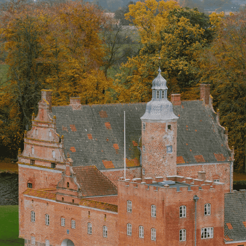Et slot i røde mursten med et enkelt tårn og efterårsfarvede træer bagved