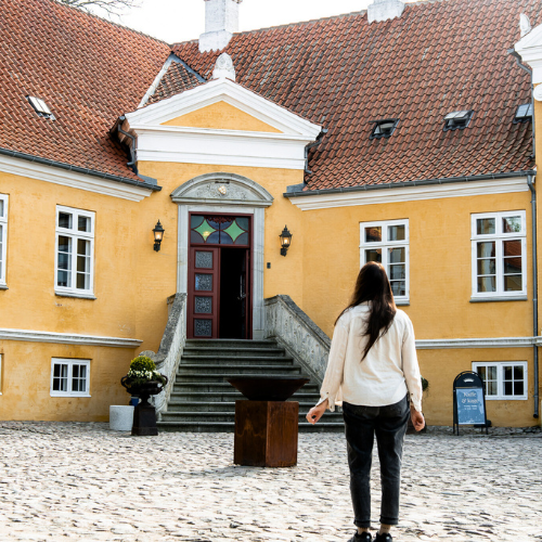 En langhåret kvinde står foran en gul herregård med ryggen til