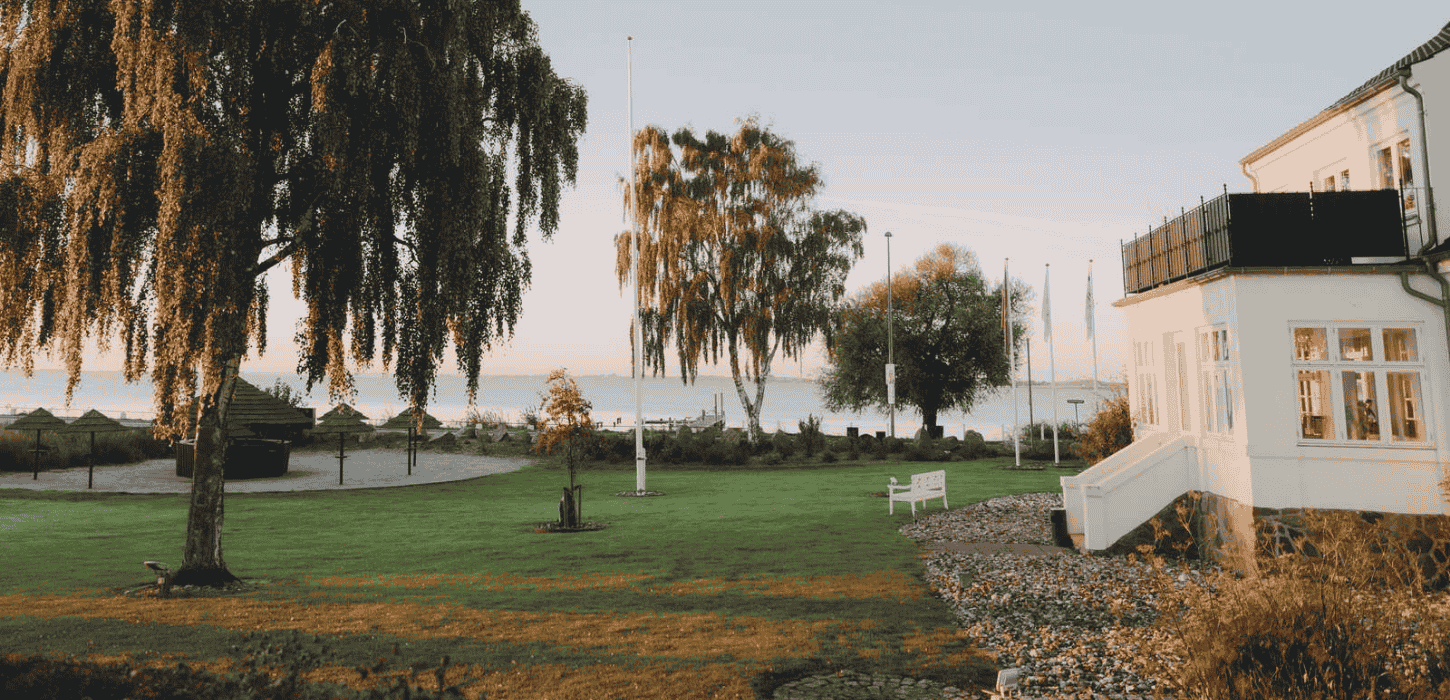 En hvid bygning med havudsigt og store piletræer foran på en græsplæne