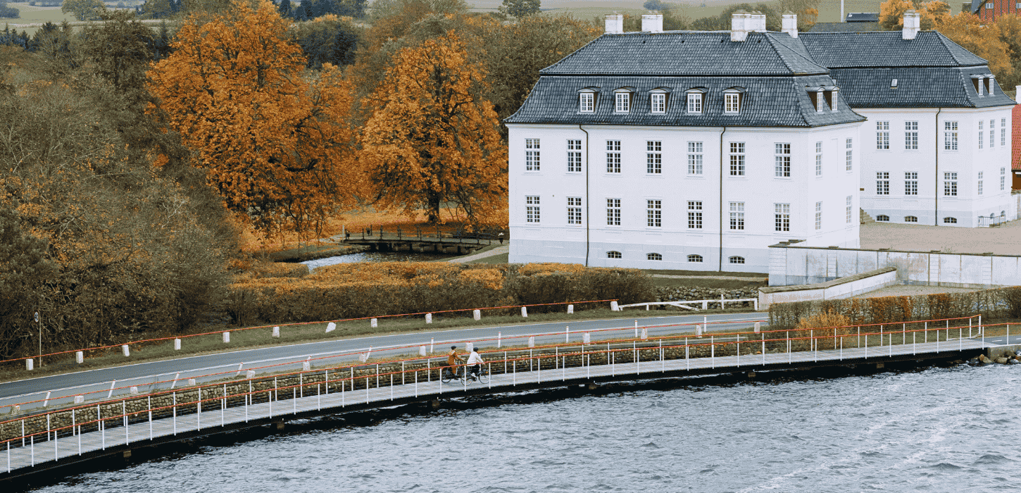 To cyklister kører på en cykelsti ude på vandet foran et hvidt gods en overskyet dag