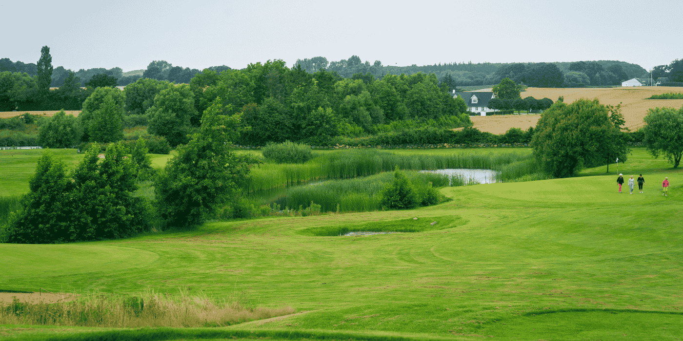 Tre golfspillere går på en bølgende og bakkende golfbane med grønne træer og marker i horisonten