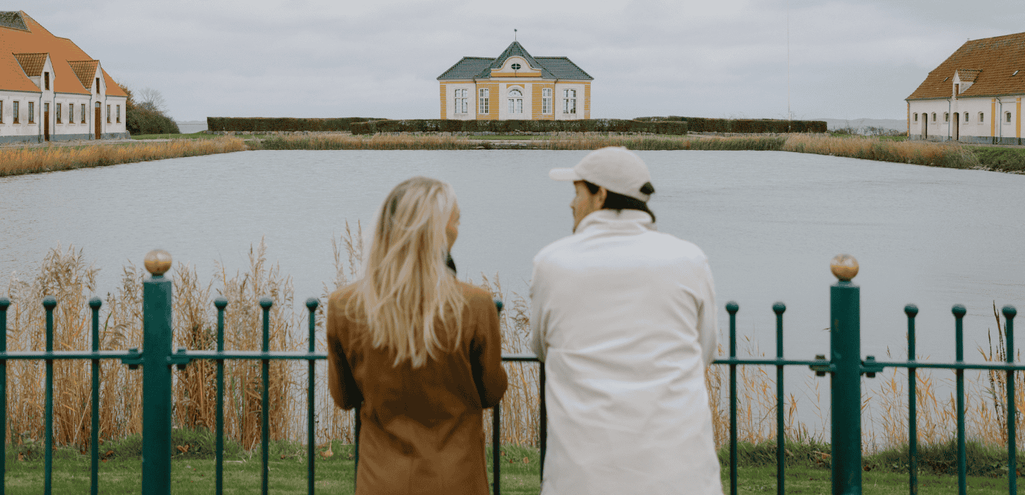 En lyshåret kvinde og mørkhåret mand står side om side med ryggen til foran et gult slot med en sø foran