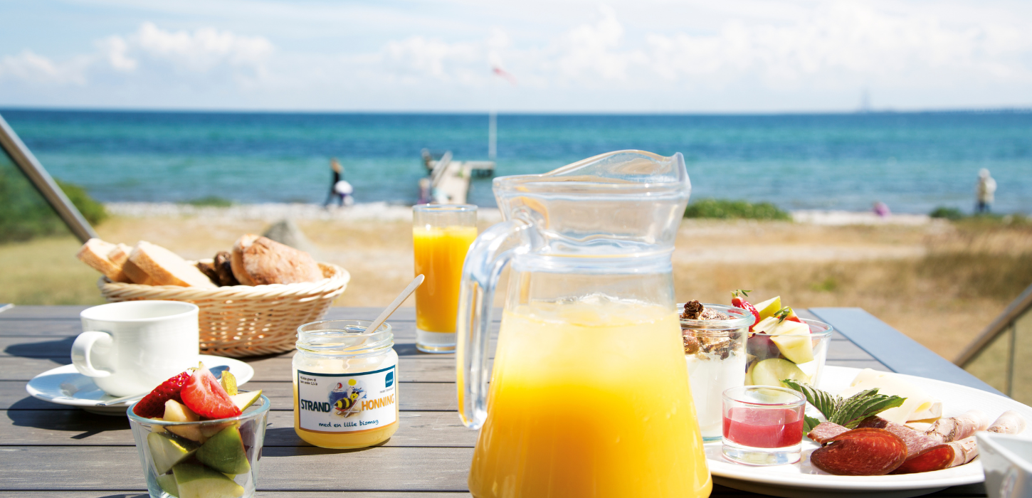 Juice, honning og andre lækre morgenmadsprodukter i forgrunden,. Strand, hav og blå himmel i baggrunden.
