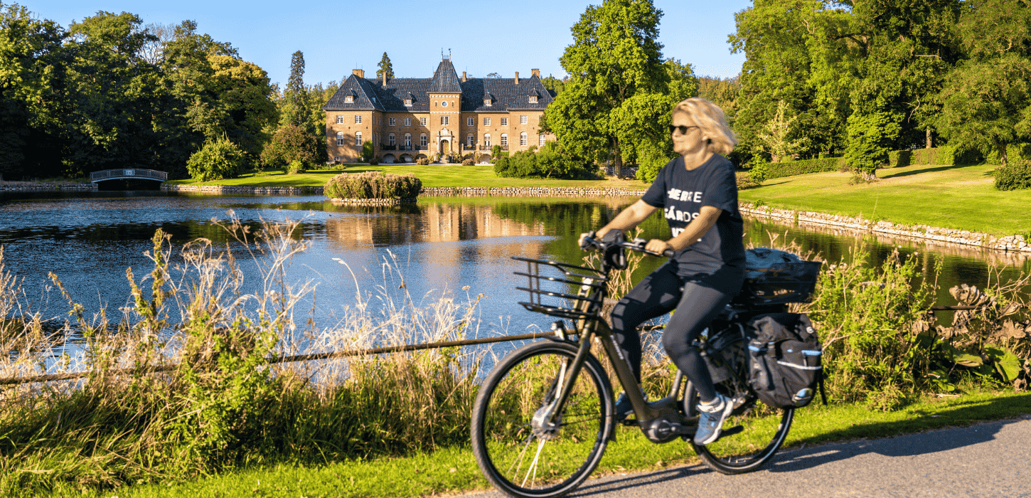 En kvinde cykler ved en sø foran et gods, omgivet af natur og sollys, nyder en fredelig dag udendørs.