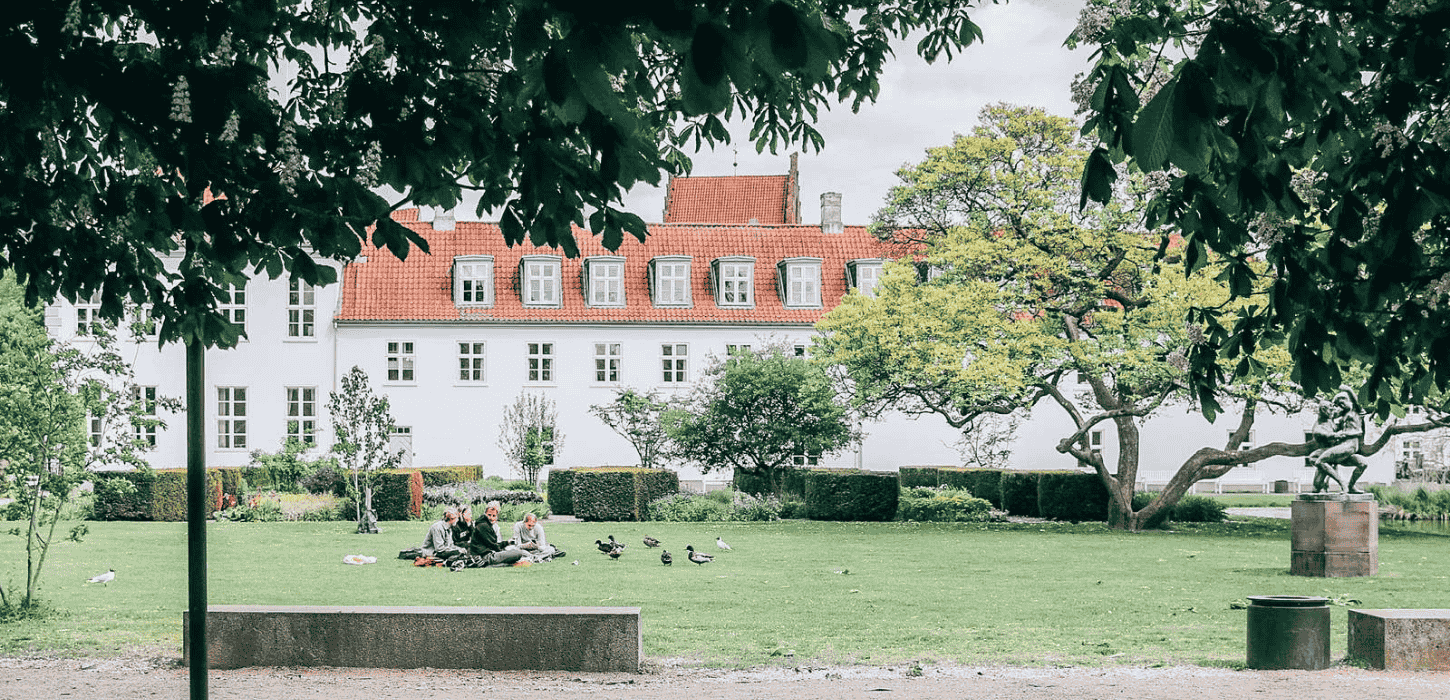 En grøn græsplæne i en park hvor der sidder en gruppe mennesker på et picnictæppe foran et hvidt gods