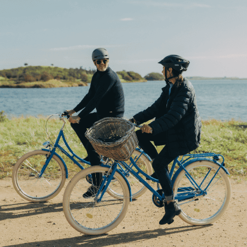 En mand og kvinde på hver deres blå cykel iført efterårsbeklædning cykler lige ud til vandet på en grussti