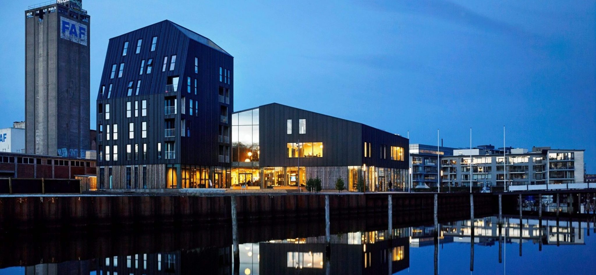 En klynge af blandede bygninger nede ved en havnefront om aftenen med tændte lys