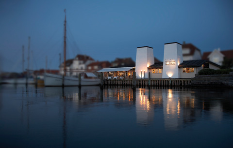 Hvid facade ud til en havn, hvor der ligger skibe foran, og det er aften med tændte lys