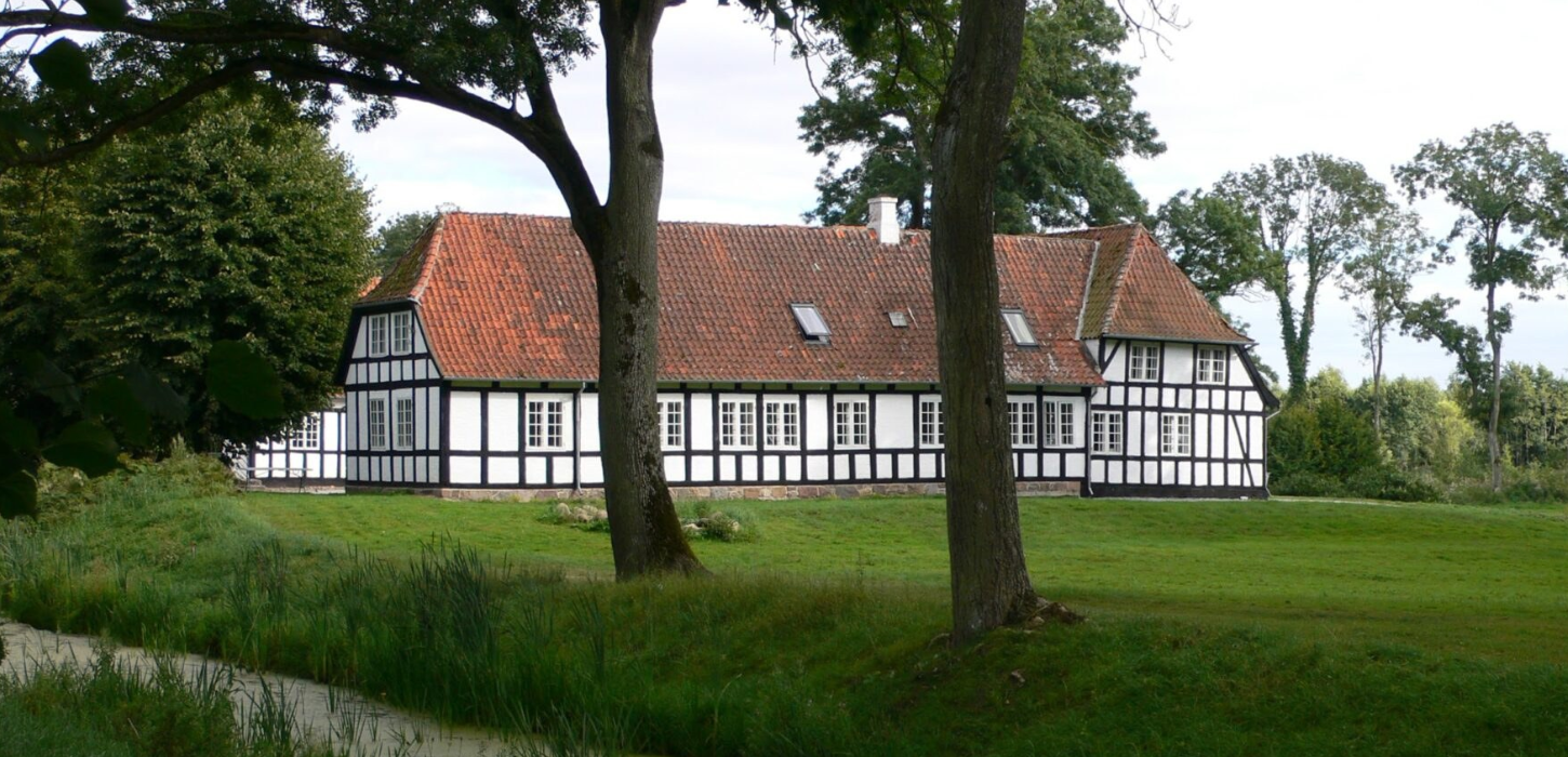 En herregård i hvidt bindingsværk med rødt tag, med et stort træ foran, der fremhæver ejendommens charme og naturlige omgivelser.