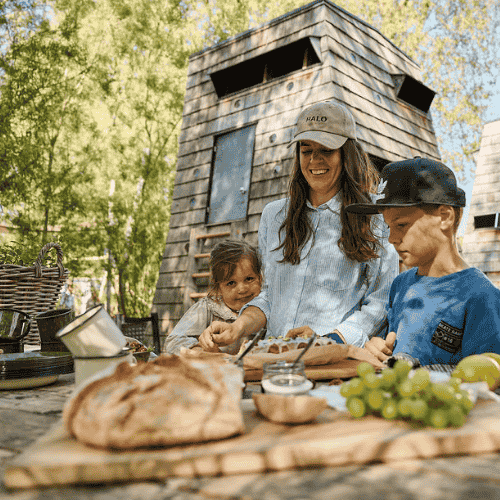 En familie bestående af en mor, en datter og en søn, iført sommertøj, laver mad udendørs foran et shelter