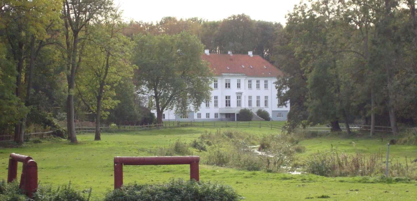 Flot hvid herregård med rødt tag ses i baggrunden af en stor grøn græsfold i forgunden.