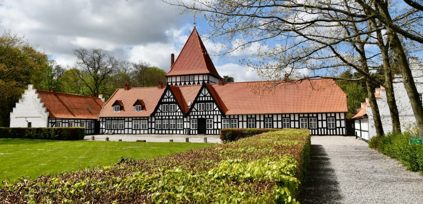 Flot hvid herregård med rødt tag og spir. En lille grusvej leder op til gården, og der er en grøn plæne i forgrunden.