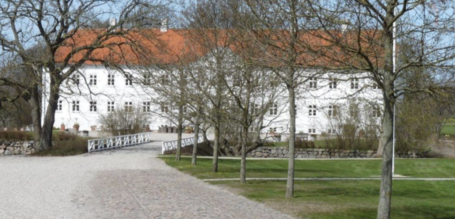 Stor hvid herregård med rødt tag og en grusvej omkranset af træer, der fører op til gården.