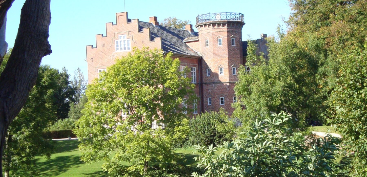 Flot rød herregård med sort tag og et tårn, omkranset af grønt græs og træer.