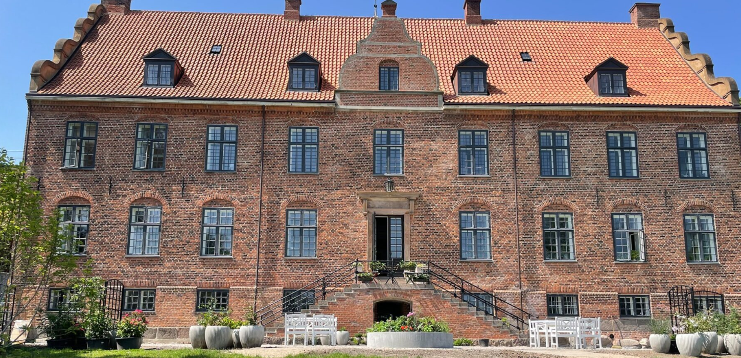 Stor rød herregård med rødt tag og grønne planter i forgrunden.