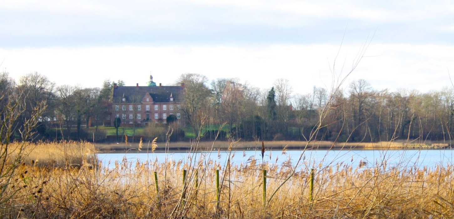 Panoramabillede af en flot sø med en stor rød herregård i baggrunden.