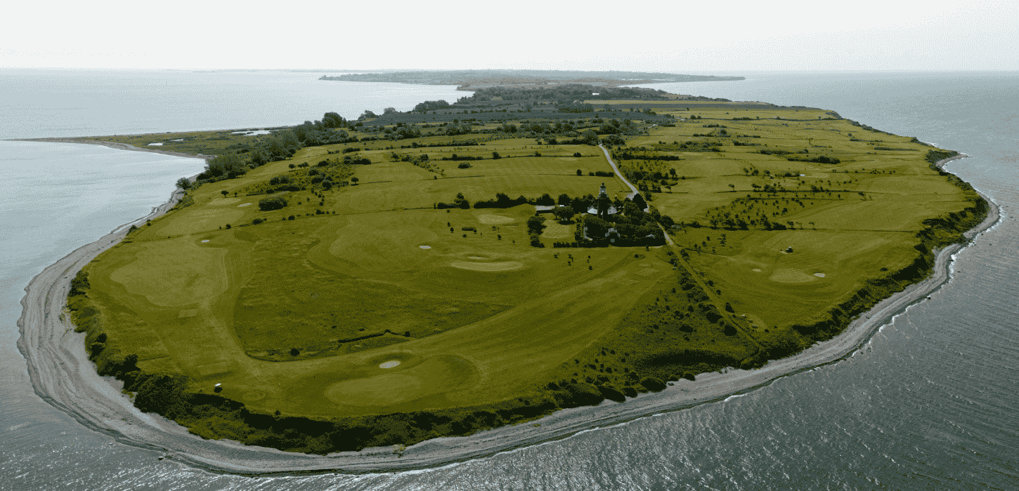 Dronebillede af en ø med en golfbane, der går helt ud til skrænten på en kyst og med et fyrtårn inde i midten