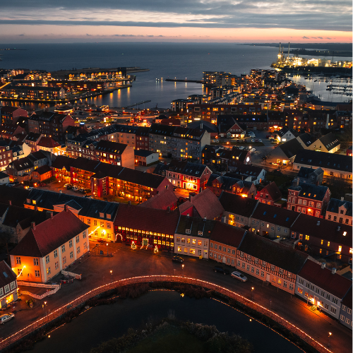 Dronefoto af Nyborg med sø og hav på hver sin side, gadebelysning og et skumringslys i horisonten