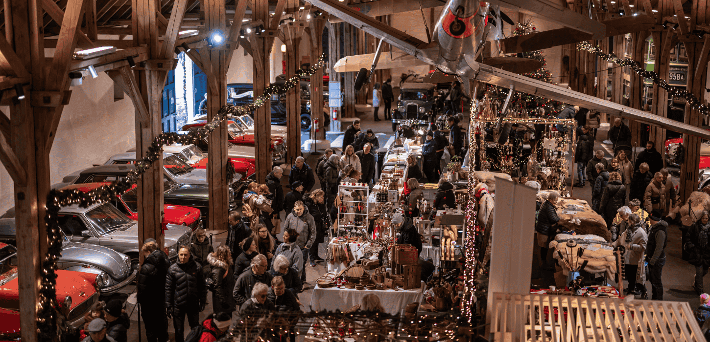 Billedet viser et travlt julemarked i Egeskov på Fyn med et festligt indendørs marked, hvor mange besøgende udforsker de forskellige boder. Markedet er pyntet med julepynt, herunder lysguirlander. Klassiske biler står opstillet langs siderne og tilføjer en unik stemning til den festlige atmosfære. Egeskov danner en livlig ramme om julen på Fyn.