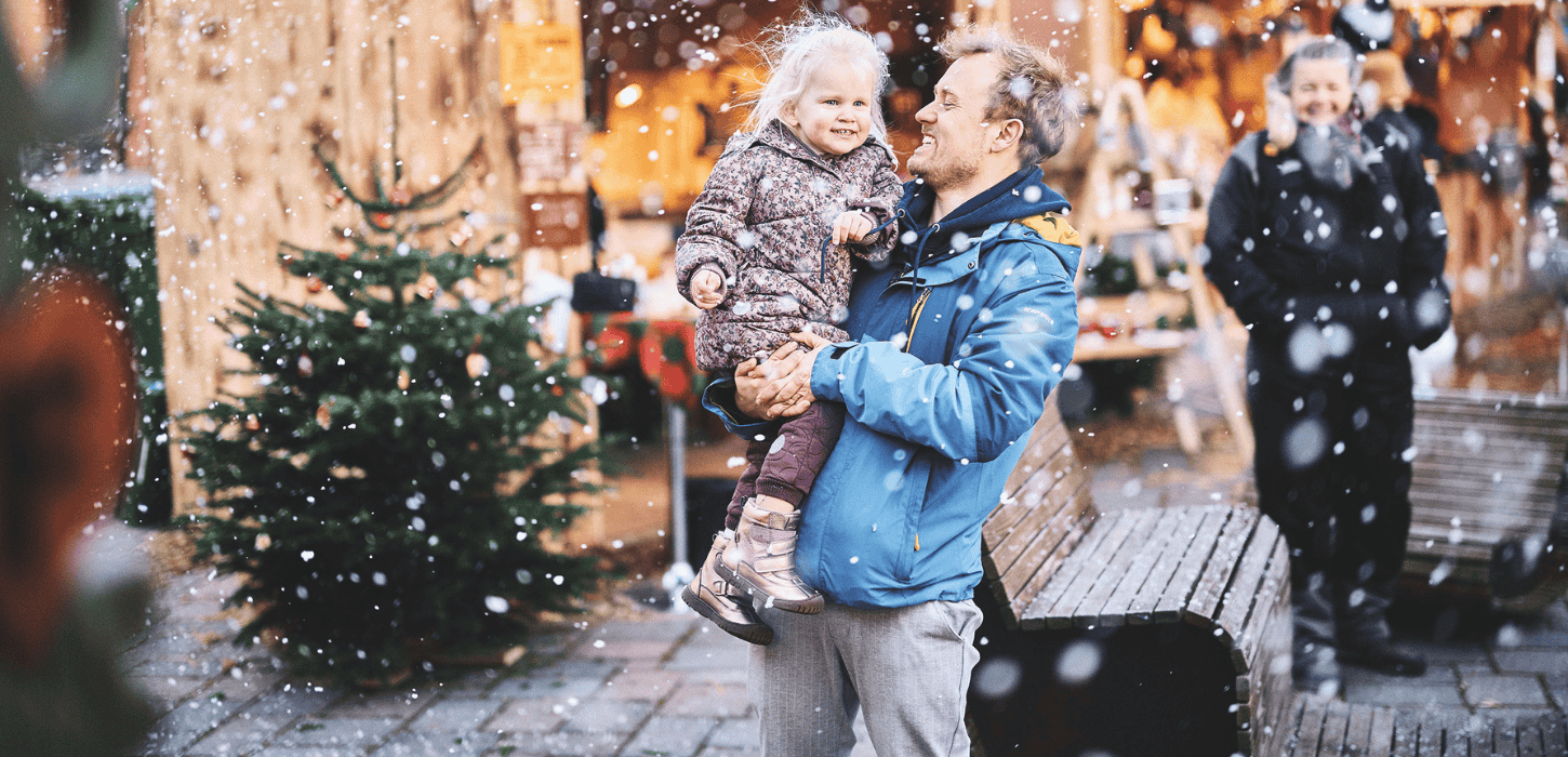 En mand holder et smilende barn i sine arme, mens han står udenfor i sneen, tæt på et juletræ og festlige julemarkedsboder.