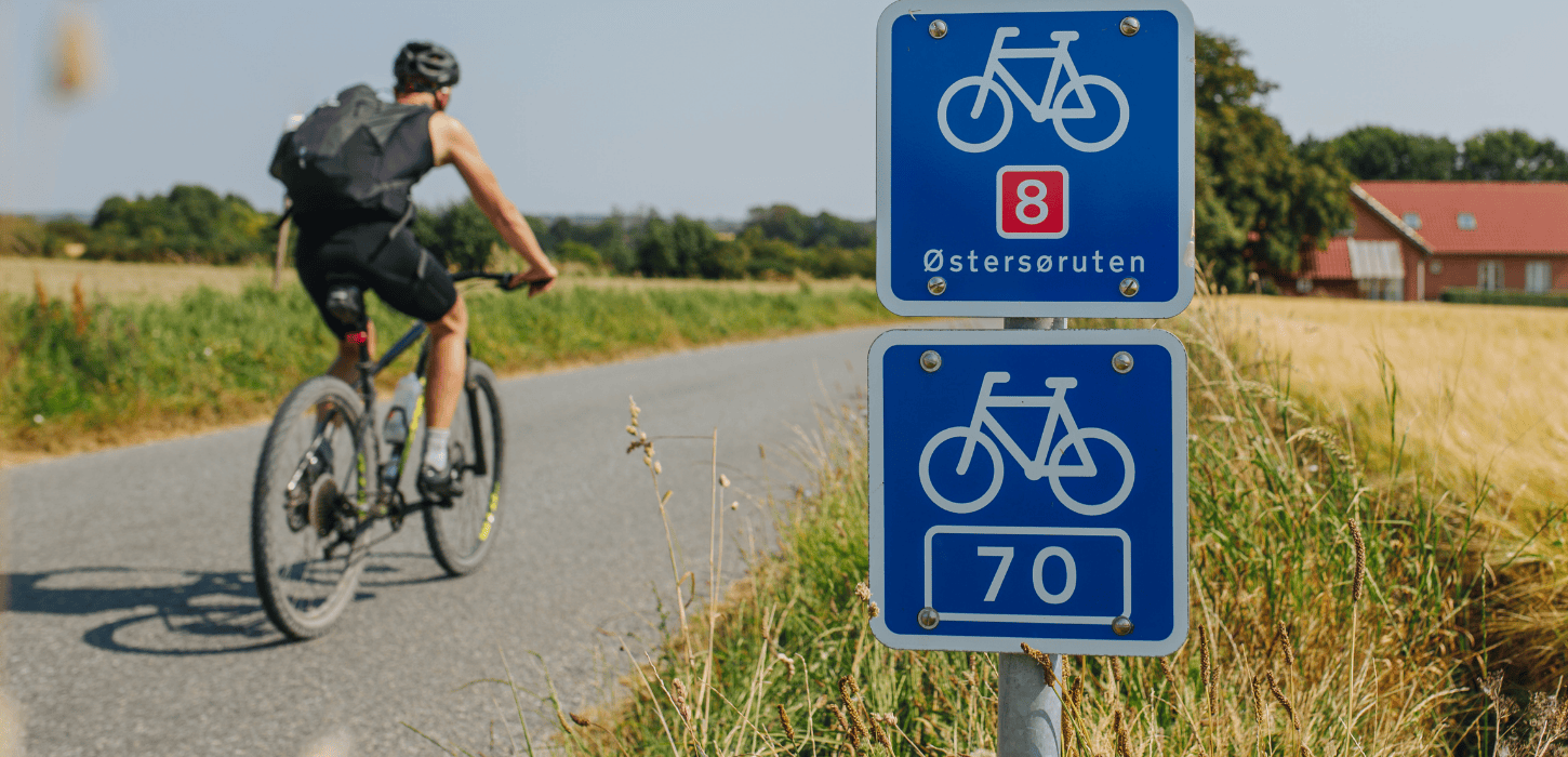 En cyklist cykler på en asfaltvej en sommerdag og i forgrunden er et blot trafikskilt med 8 tal og Østersøruten på