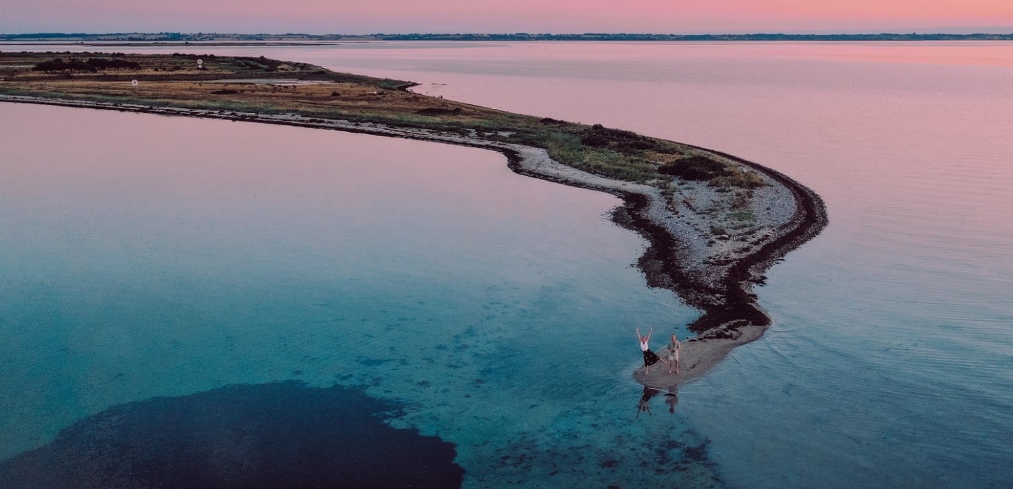 Et par står for enden af en landtange på et dronebillede med solnedgangen i baggrunden